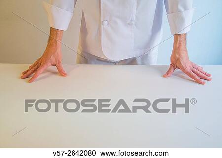 Stock Image - Cook behind a white table, hands on the table.. Fotosearch