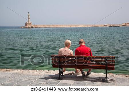 Stock Image - Couple sitting on a bench with the lighthouse in the background at the Venetian harbor of Chania, Crete, Greek Islands, Greece.. Fotosearch
