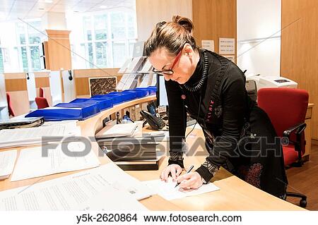 Picture - European Parliament, Brussels, Belgium. Female press officer signing a legal, while standing form behind her desk.. Fotosearch