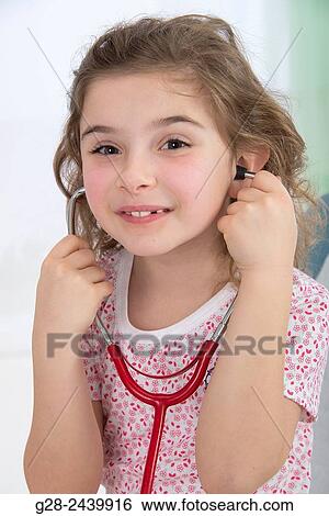 Stock Photograph - Little smiling girl sitting on a medical couch and listening to her heart with a stethoscope. Fotosearch