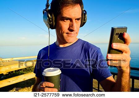 Man with blue t-shirt is listening music with his headphones on while he is using a mobile phone and is holding a plastic cup of coffee outdoor near the sea. View Large Photo Image Stock Photo - Man with blue t-shirt is listening music with his headphones on while he is using a mobile phone and is holding a plastic cup of coffee outdoor near the sea.. Fotosearch