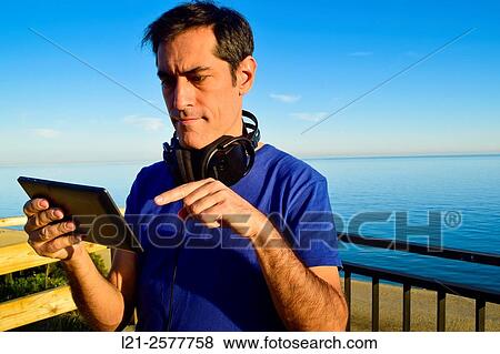 Stock Photo - Man with headphones around neck is using a tablet outdoor near the sea. He is wearing a blue t-shirt.. Fotosearch