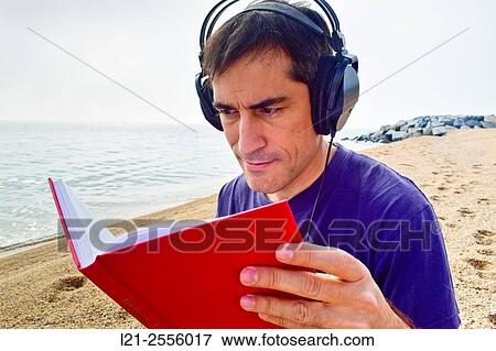 Man with headphones listening to music while he is reading a book on a beach, near the sea. View Large Photo Image Stock Photo - Man with headphones listening to music while he is reading a book on a beach, near the sea.. Fotosearch