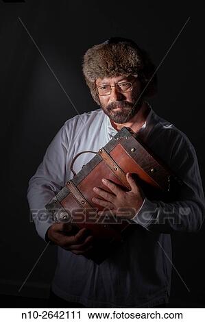 Stock Image - Middle age man, with a beard, glasses, and fur hat, holding a wooden case.. Fotosearch