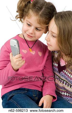 Stock Image - Pair of curious girls look a mobile phone, the younger is sitting on the knees of her friend, she has pigtails. Fotosearch