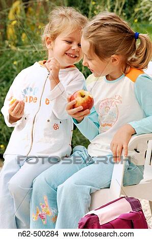 Pair of girls eating an apple outdoors, a little girl is sitting on the chair while the other is standing next to her View Large Photo Image Picture - Pair of girls eating an apple outdoors, a little girl is sitting on the chair while the other is standing next to her. Fotosearch