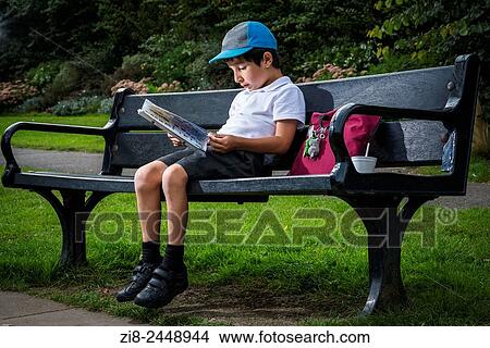 Primary schoolboy sitting on a park bench reading a book, Surrey, UK. View Large Photo Image Picture - Primary schoolboy sitting on a park bench reading a book, Surrey, UK.. Fotosearch