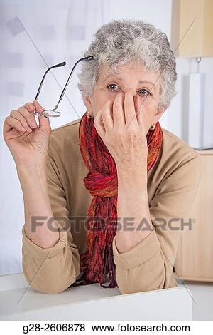 Stock Photo - Senior woman with grey hair, holding top of her nose with one hand to relieve sinusitis and headache, with one hand the other hand is holding her glasses. Fotosearch