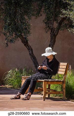 Senior woman with sun hat, sitting on a public wooden bench, reading her tablet, in a garden, Marrakech, Morocco View Large Photo Image Stock Image - Senior woman with sun hat, sitting on a public wooden bench, reading her tablet, in a garden, Marrakech, Morocco. Fotosearch
