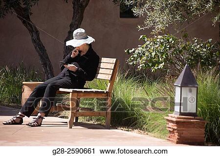 Senior woman with sun hat, sitting on a public wooden bench, reading her tablet, in a garden, Marrakech, Morocco View Large Photo Image Stock Image - Senior woman with sun hat, sitting on a public wooden bench, reading her tablet, in a garden, Marrakech, Morocco. Fotosearch