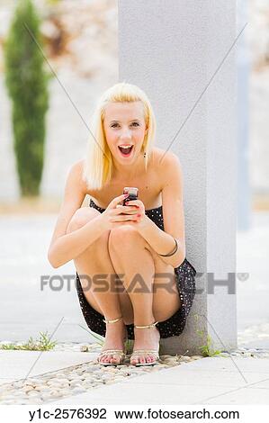 Stock Image - Teen girl squatting with cellular phone in hands is surprised. Fotosearch