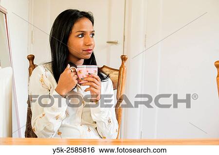 Stock Photograph - Tilburg, Netherlands. Young, dark colored woman drinking a warm cup of thee, while sitting at her kitchen table.. Fotosearch