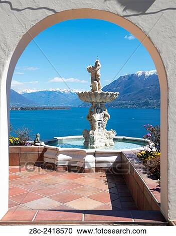 Stock Image - Window view with a water fountain with statue and panoramic view over alpine lake Maggiore with snow-capped mountains in a sunny day with blue sky in Ticino, Switzerland.. Fotosearch