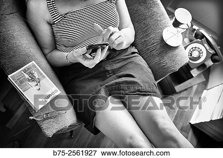 Stock Photo - Woman sitting in a armchair, in a relax position, handling a mobile phone in a town house. Mahn, Menorca, Balearic Islands, Spain, Europe.. Fotosearch