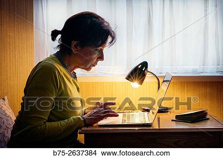 Picture - woman working with a laptop, with glasses and a lighted lamp on the table.. Fotosearch