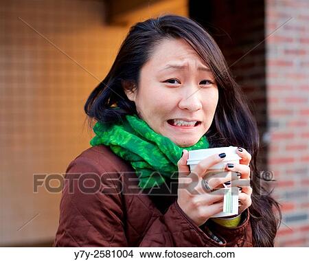 Young Asian woman makes a face while warming her hands with tea in a paper cup in the winter streets of Manhattan. View Large Photo Image Picture - Young Asian woman makes a face while warming her hands with tea in a paper cup in the winter streets of Manhattan.. Fotosearch