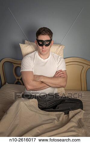 Stock Photo - Young man, sitting on bed, wearing black mask.. Fotosearch