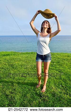 Young woman in summer clothes and straw hat standing on the grass near the sea. She is dressed with short jeans and a white blouse. View Large Photo Image Stock Image - Young woman in summer clothes and straw hat standing on the grass near the sea. She is dressed with short jeans and a white blouse.. Fotosearch