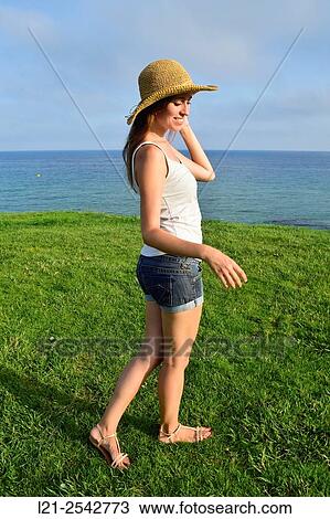 Stock Image - Young woman in summer clothes and straw hat standing on the grass near the sea. She is dressed with short jeans and a white blouse.. Fotosearch