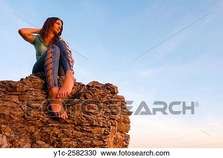 Stock Image - Young woman is watching sunset sitting atop big rock. Fotosearch