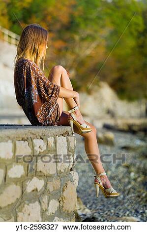 Stock Photo - Young woman sitting on wall atop beach watching sunset. Fotosearch