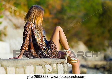 Stock Photo - Young woman sitting on wall atop beach watching sunset. Fotosearch
