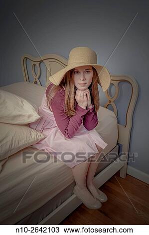 Stock Image - Young woman, wearing a hat, sitting on a bed.. Fotosearch