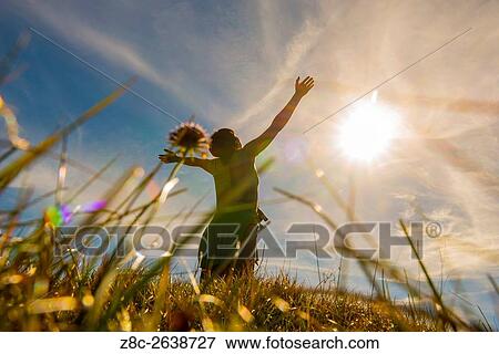 Stock Photo - Low angle view on a woman with raised arms and grass and sunshine in Grisons, Switzerland.. Fotosearch