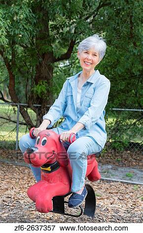 Retired woman in her 70s at playground playfully riding toy dog Clifford the Big Red Dog Model Released MR-2. View Large Photo Image Stock Photo - Retired woman in her 70s at playground playfully riding toy dog Clifford the Big Red Dog Model Released MR-2.. Fotosearch