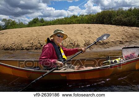 Stock Photograph - Senior man with sunglasses and hat solo paddling a small canoe near a sandy beach on a tranquil Moose River in Old Forge, New York.. Fotosearch