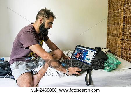 Stock Photo - Tilburg, Netherlands. Brazilian male traveller sitting on the bed of his Airbnb guest room with his laptop computer at arms length.. Fotosearch