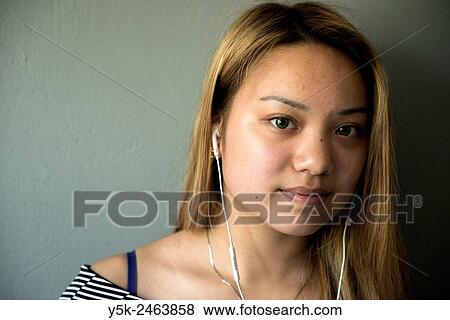 Stock Photo - Tilburg, Netherlands. Studio portrait of a young Chinese girl with headphones, listening to the music on her smartphone, while standing in front of a grey background.. Fotosearch