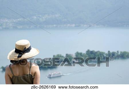 Stock Photo - Woman with a straw hat watching Brissago islands and passenger ship on alpine lake Maggiore in Ticino, Switzerland.. Fotosearch
