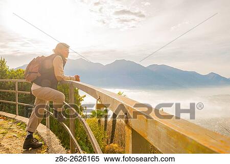 Stock Photography - Woman with backpack leaning on a railing and enjoy panoramic view with sea of fog over alpine lake Maggiore with mountain in Ticino, Switzerland.. Fotosearch