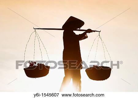 Stock Photo - Woman with conical hat carries flowers in pannier baskets in blazing sun across white sand dunes near Mui Ne Vietnam. Fotosearch