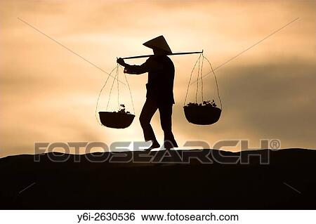 Stock Photograph - Woman with conical hat carries flowers in pannier baskets over sand dunes at sunrise near Mui Ne Vietnam.. Fotosearch