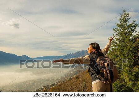 Picture - Woman with outstretched arms and backpack enjoy panoramic view with sea of fog over alpine lake Maggiore with mountain in Ticino, Switzerland.. Fotosearch