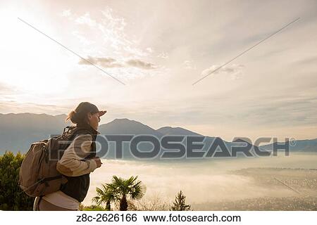 Stock Photograph - Woman with raised hand and backpack enjoy panoramic view with sea of fog over alpine lake Maggiore with mountain in Ticino, Switzerland.. Fotosearch