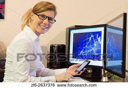 Stock Photo - Young woman working at home office using cell phone and two computer monitors doing business with clients on the internet MR Model released.. Fotosearch