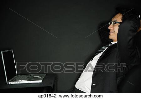 Businessman leaning back, facing laptop, smiling View Large Photo Image Stock Image - Businessman leaning back, facing laptop, smiling. Fotosearch