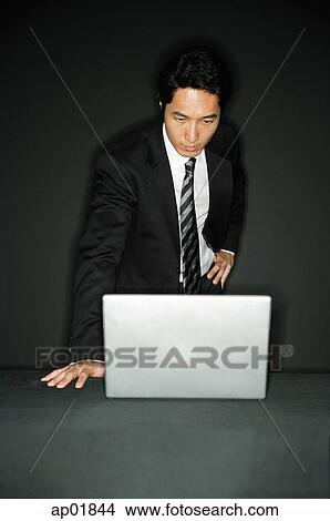 Picture - Businessman standing, looking at laptop on table. Fotosearch