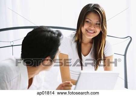 Picture - Couple in bedroom, woman smiling, man looking at her, laptop in front of her. Fotosearch