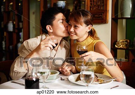 Stock Image - Couple in Chinese restaurant, holding wine glasses, man kissing woman on cheek. Fotosearch