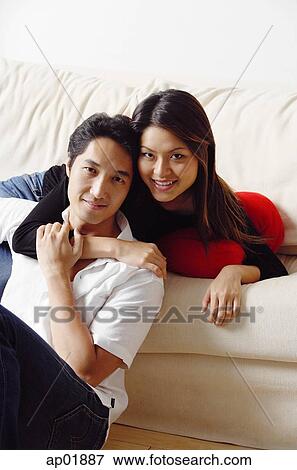 Stock Photo - Couple in living room, woman lying in sofa, arm around man sitting in front of her. Fotosearch