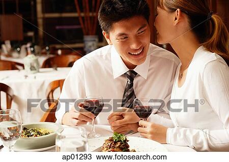 Stock Image - Couple in restaurant, sitting side by side, woman whispering into man's ear. Fotosearch