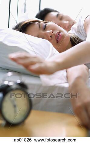 Stock Photograph - Couple lying on bed, woman reaching to switch of alarm clock in foreground. Fotosearch