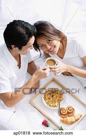 Stock Photo - Couple on bed, breakfast tray between them, woman looking at camera. Fotosearch