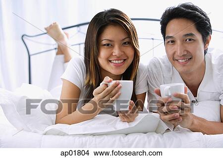 Couple on bed, holding mugs, smiling at camera, portrait View Large Photo Image Picture - Couple on bed, holding mugs, smiling at camera, portrait. Fotosearch