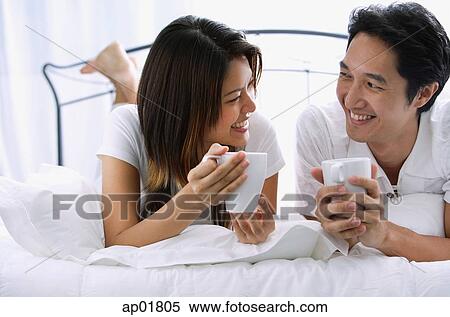 Stock Photography - Couple on bed, holding mugs, smiling at each other. Fotosearch