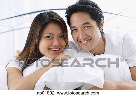 Stock Image - Couple on bed, hugging pillows, smiling at camera, portrait. Fotosearch